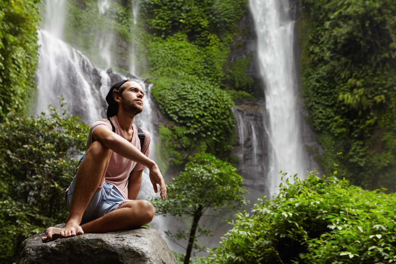 Attractive young bearded adventurer wearing no shoes having break on big rock while trekking alone in tropical forest. Stylish hiker relaxing outdoors in jungle with amazing waterfall in background